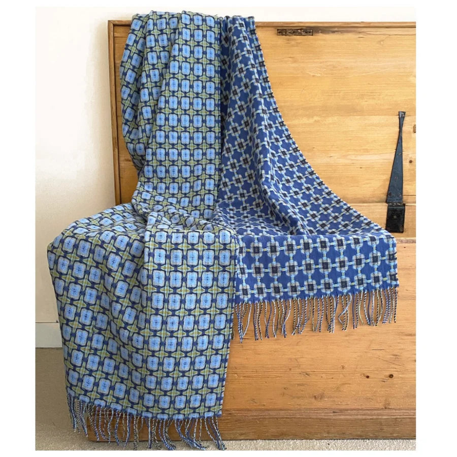 Reversible woven throw blanket with blue geometric patterns on each side and knotted fringe trim draped over wooden bench