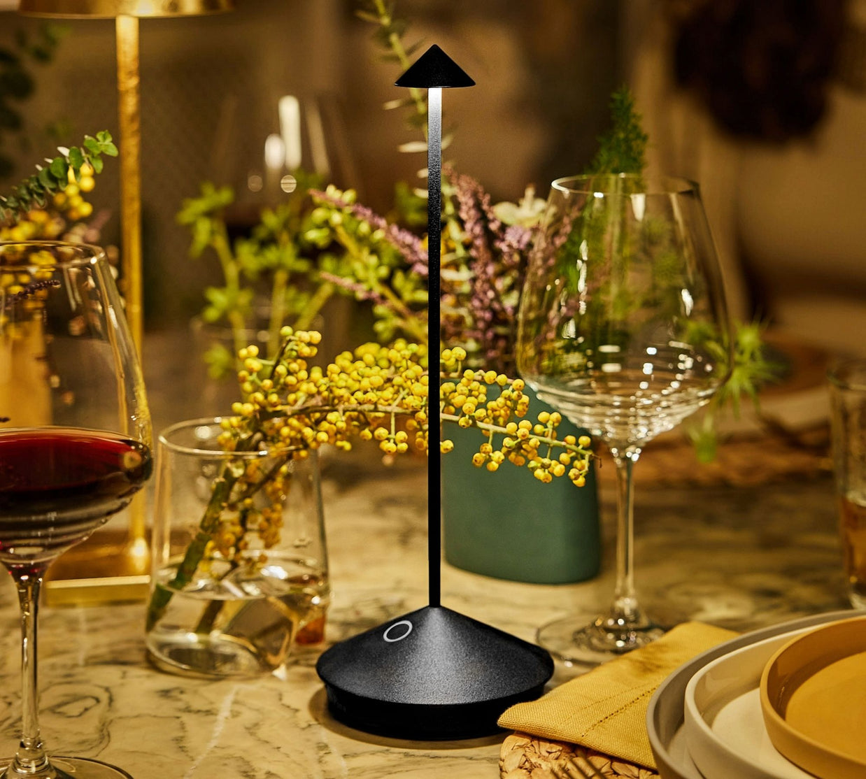 Modern black triangular LED table lamp with slender stem and circular base on dining table with wine glasses and flowers